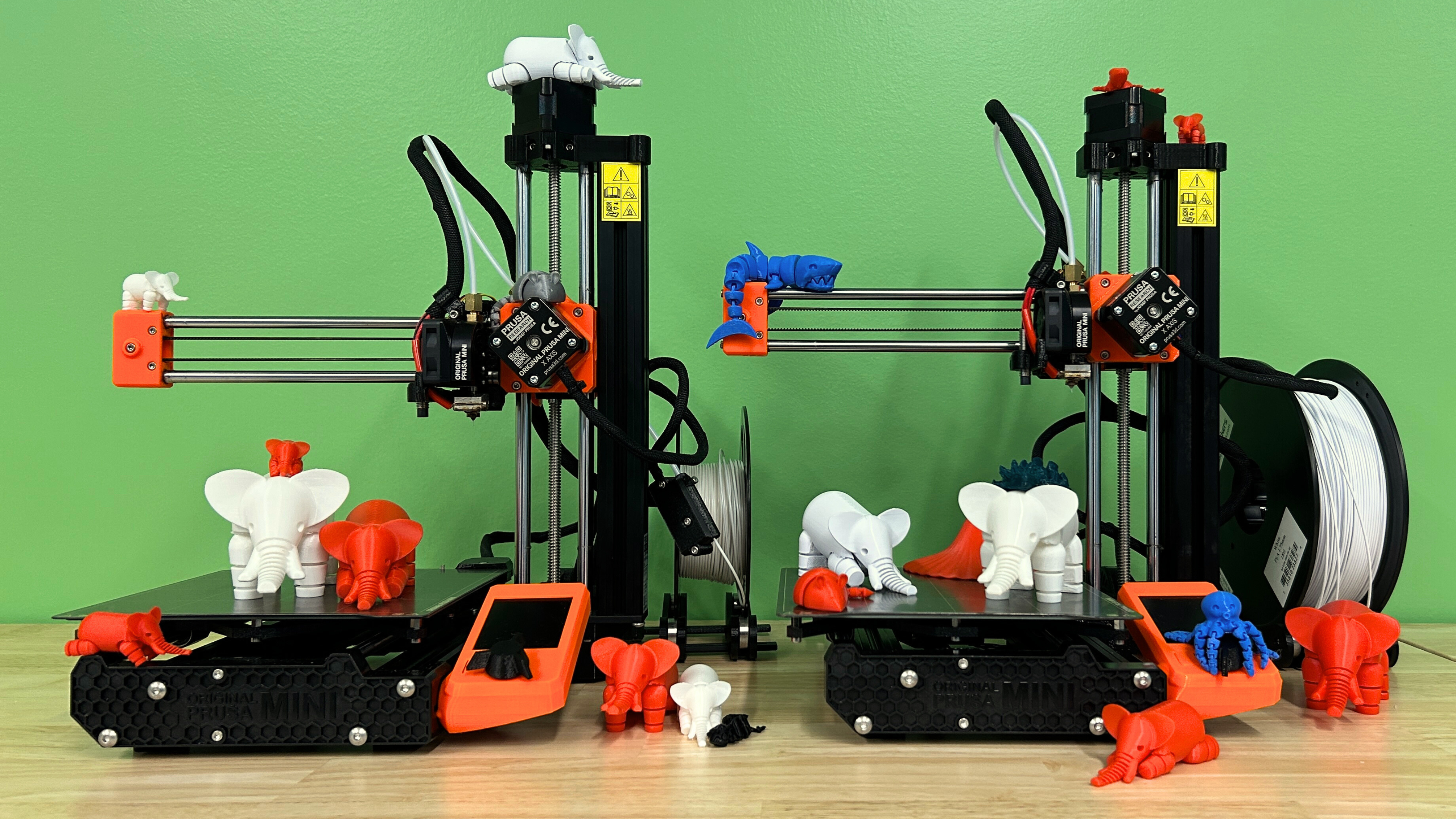Two Prusa Mini 3D printers against a green background with various 3D printed animals perched all over them!